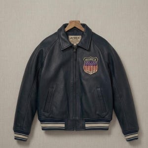 Op de afbeelding: Een donker marineblauw leren bomberjack met een ritssluiting aan de voorzijde, een omgeslagen kraag en geribbelde boorden en zoom met witte strepen. Een schildvormige patch op de borst toont de tekst AVIREX en een vlagafbeelding met sterren en strepen.