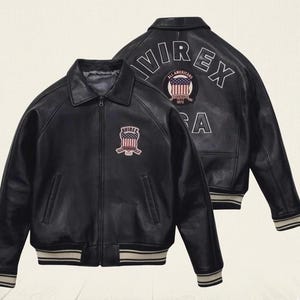 Puede incluir: Una chaqueta de cuero negra con cremallera frontal, cuello y pu&ntilde;os y dobladillo de canal&eacute; a rayas. El frente muestra un parche con la bandera estadounidense y el texto AVIREX. La parte posterior presenta el texto AVIREX USA y un parche circular.