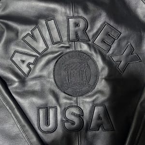 Puede incluir: Una chaqueta de cuero negra con la palabra AVIREX arqueada en la parte superior en letras grandes, negritas y cosidas. Debajo, un parche circular muestra el texto ALL AMERICAN ESTABLISHED 1975. La palabra USA est&aacute; cosida en letras grandes abajo.