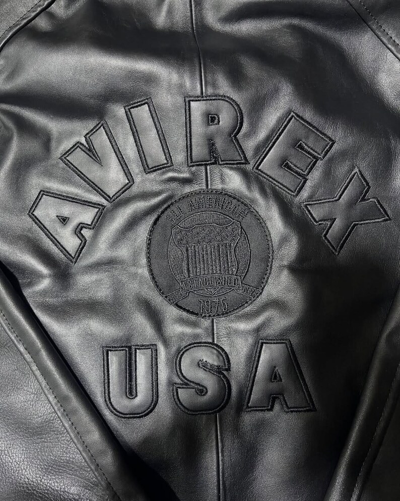 Puede incluir: Una chaqueta de cuero negra con la palabra AVIREX arqueada en la parte superior en letras grandes, negritas y cosidas. Debajo, un parche circular muestra el texto ALL AMERICAN ESTABLISHED 1975. La palabra USA est&aacute; cosida en letras grandes abajo.