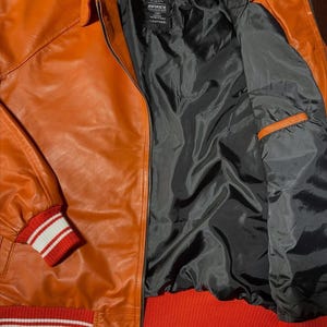 Op de afbeelding: Een oranje leren jas met een zwarte voering en een zwart merklabel met de tekst AVIREX. De jas is voorzien van een ritssluiting aan de voorzijde, een klassieke kraag en geribbelde gebreide boorden aan de mouwen en zoom in een rood-wit gestreept patroon.