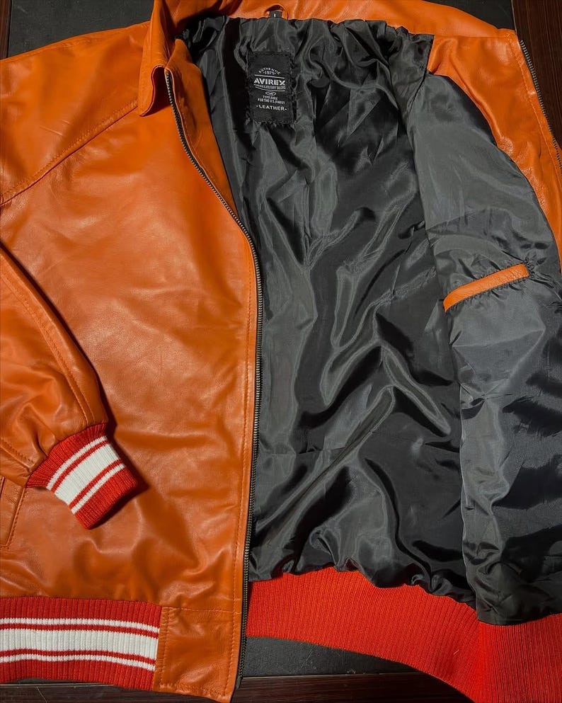 Op de afbeelding: Een oranje leren jas met een zwarte voering en een zwart merklabel met de tekst AVIREX. De jas is voorzien van een ritssluiting aan de voorzijde, een klassieke kraag en geribbelde gebreide boorden aan de mouwen en zoom in een rood-wit gestreept patroon.
