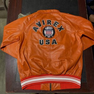 Op de afbeelding: Een oranje leren jas met een geribbelde zoom met witte en rode strepen. Op de achterkant staat de tekst AVIREX en USA in zwarte letters, samen met een ronde badge met een schild, sterren en de tekst ALL AMERICANS ESTABLISHED 1975.