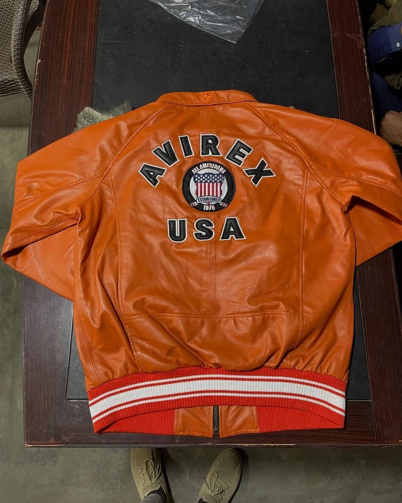 Op de afbeelding: Een oranje leren jas met een geribbelde zoom met witte en rode strepen. Op de achterkant staat de tekst AVIREX en USA in zwarte letters, samen met een ronde badge met een schild, sterren en de tekst ALL AMERICANS ESTABLISHED 1975.