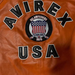 Op de afbeelding: Een oranje leren jas met de zwart geborduurde tekst AVIREX en USA. Een ronde patch in het midden toont een schild met sterren en strepen, de tekst ALL AMERICANS, ESTABLISHED en 1975, met N.Y. aan de zijkanten.