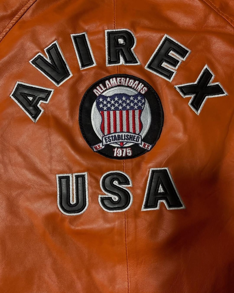 Op de afbeelding: Een oranje leren jas met de zwart geborduurde tekst AVIREX en USA. Een ronde patch in het midden toont een schild met sterren en strepen, de tekst ALL AMERICANS, ESTABLISHED en 1975, met N.Y. aan de zijkanten.