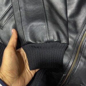 Puede incluir: Una chaqueta de cuero negro con un pu&ntilde;o de punto acanalado en el extremo de la manga, detalles de costuras verticales a lo largo del brazo y un cierre de cremallera met&aacute;lico en la parte delantera. La chaqueta tiene una textura lisa.