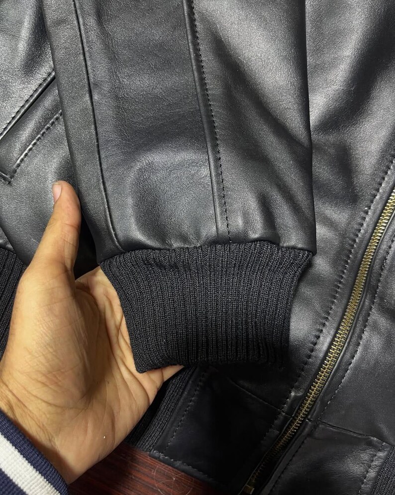 Puede incluir: Una chaqueta de cuero negro con un pu&ntilde;o de punto acanalado en el extremo de la manga, detalles de costuras verticales a lo largo del brazo y un cierre de cremallera met&aacute;lico en la parte delantera. La chaqueta tiene una textura lisa.