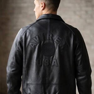 Puede incluir: Una chaqueta de cuero negra con cuello vuelto, mangas largas con pu&ntilde;os acanalados y dobladillo acanalado. La parte posterior muestra el texto en relieve "AVIREX" en arco sobre un emblema circular y la palabra "USA" centrada debajo.