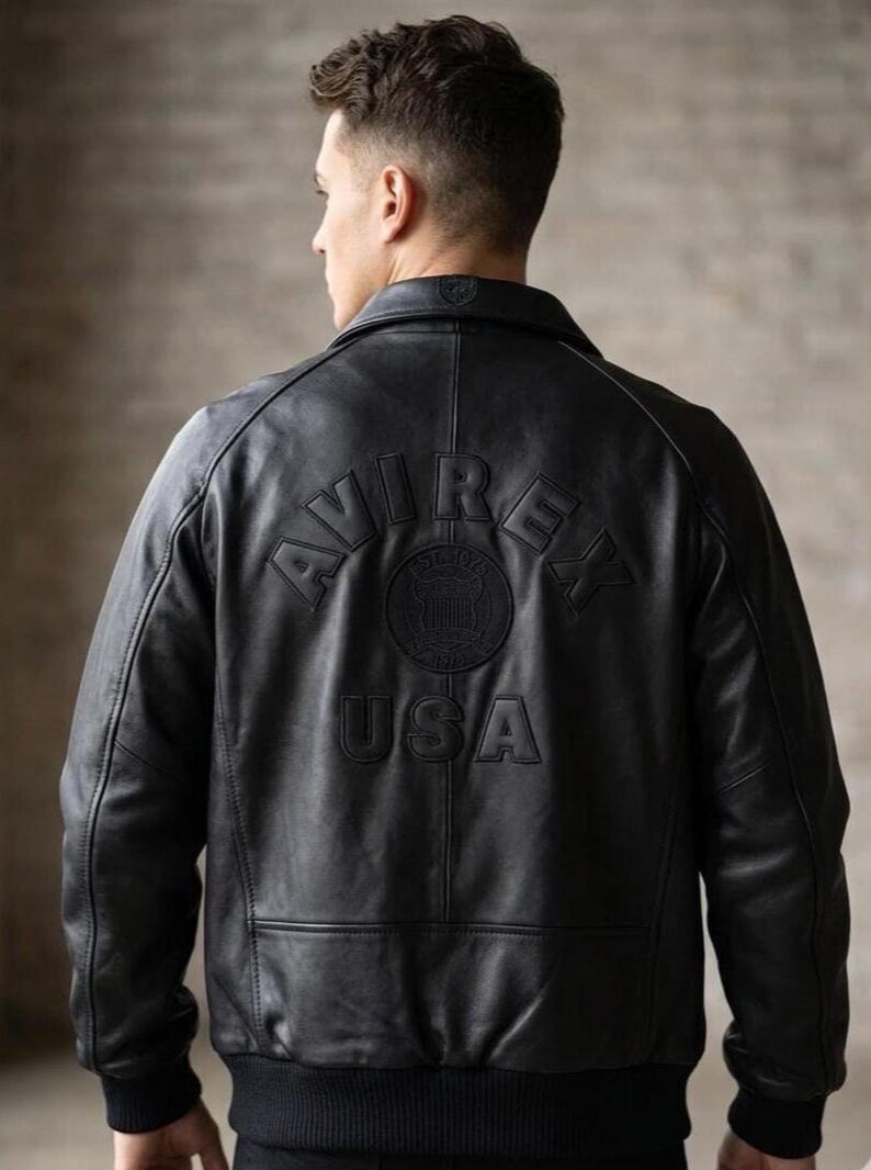 Puede incluir: Una chaqueta de cuero negra con cuello vuelto, mangas largas con pu&ntilde;os acanalados y dobladillo acanalado. La parte posterior muestra el texto en relieve "AVIREX" en arco sobre un emblema circular y la palabra "USA" centrada debajo.