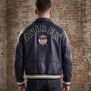 Puede incluir: Una chaqueta de cuero azul marino oscuro con el texto AVIREX arqueado en letras blancas en la parte superior de la espalda. Debajo del texto hay un parche circular que muestra una bandera estadounidense y las palabras ALL AMERICANS ESTABLISHED 1875.