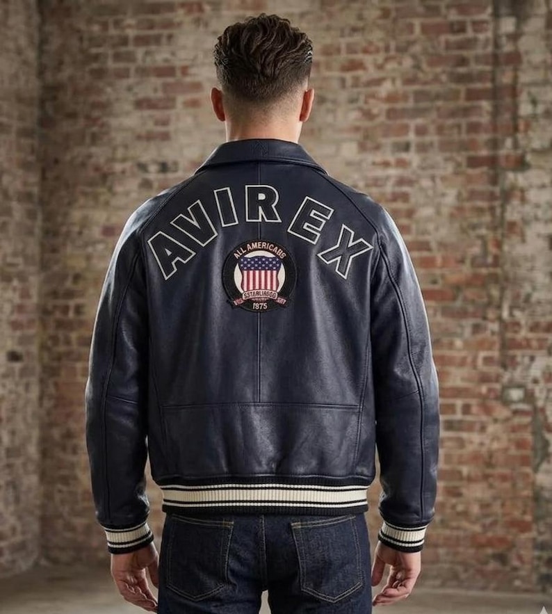 Puede incluir: Una chaqueta de cuero azul marino oscuro con el texto AVIREX arqueado en letras blancas en la parte superior de la espalda. Debajo del texto hay un parche circular que muestra una bandera estadounidense y las palabras ALL AMERICANS ESTABLISHED 1875.