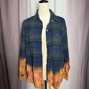 May include: A long-sleeved, button-down flannel shirt in a green, navy, and red plaid pattern. The bottom of the shirt and sleeves have been dyed orange and pink. The shirt is displayed on a mannequin.