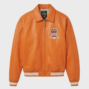 May include: Orange leather bomber jacket with a collared neckline and a zip-up front. Features a patch with an American flag design and the brand name. The cuffs and hem have white and orange striped detailing.