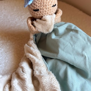 Peut inclure: Une couverture de sécurité pour bébé faite à la main avec une tête de poupée crochetée avec les yeux fermés et un petit nœud bleu. La poupée est attachée à un tissu bleu clair doux et à une matière crème en peluche.