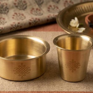 May include: A brass bowl and a matching brass tumbler, both featuring a delicate, etched geometric pattern on the exterior. The items have a smooth, metallic finish and a wide, rounded rim, showcasing a consistent design aesthetic in their construction.