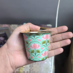 May include: A small, cylindrical metal cup featuring a light mint green background decorated with a repeating pattern of pink lotus flowers and green leaves. The cup has a smooth, metallic gold-toned rim and a tapered base design.
