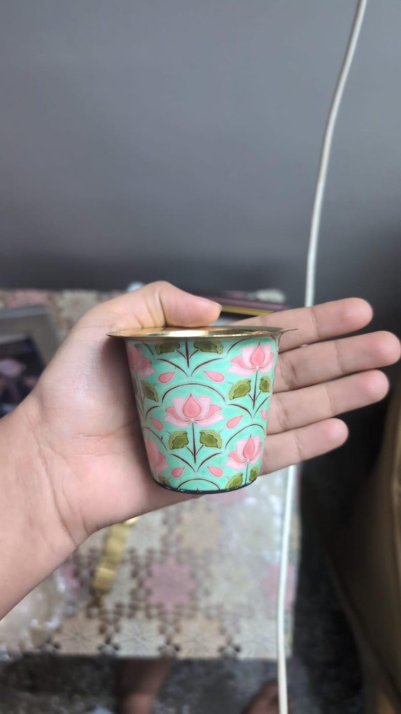 May include: A small, cylindrical metal cup featuring a light mint green background decorated with a repeating pattern of pink lotus flowers and green leaves. The cup has a smooth, metallic gold-toned rim and a tapered base design.