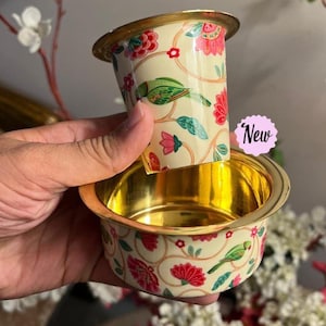May include: A two-piece metal coffee filter set with a cream-colored exterior featuring a floral and green parrot pattern. The interior of both pieces has a polished gold finish. A small pink scalloped sticker with the text New is attached.