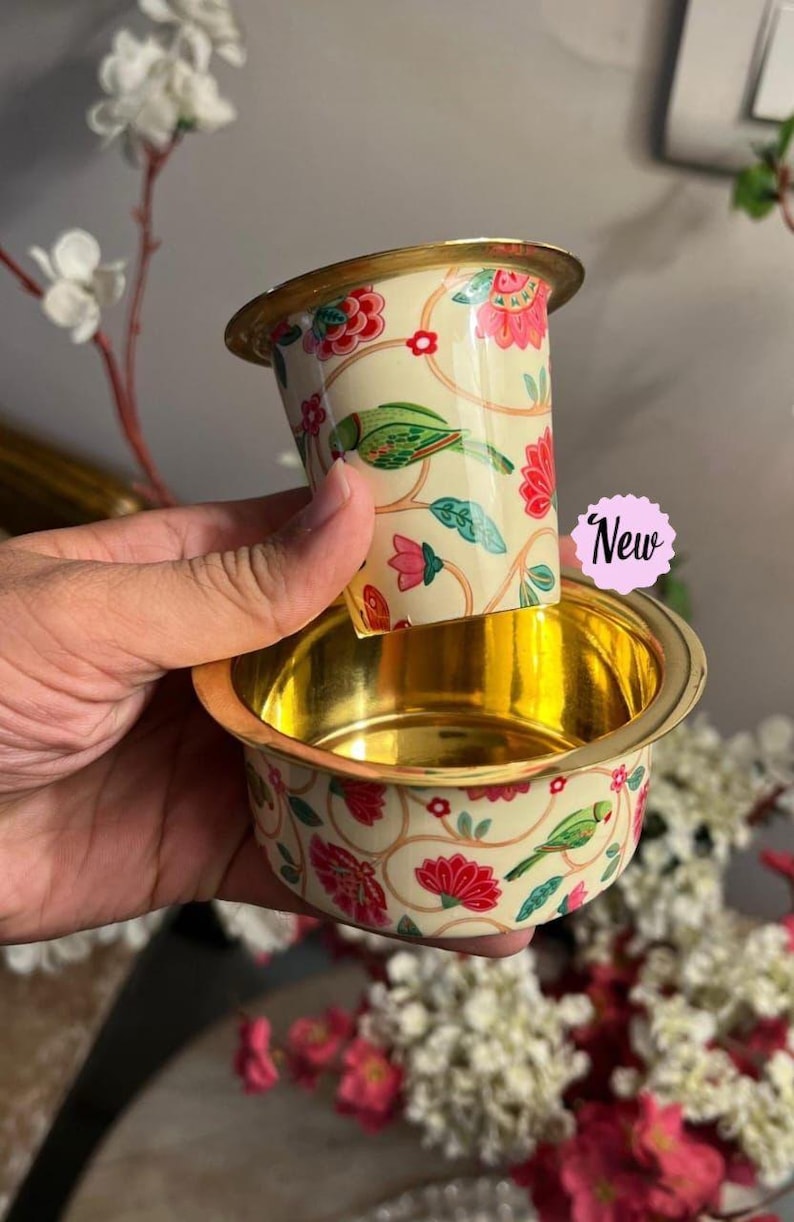 May include: A two-piece metal coffee filter set with a cream-colored exterior featuring a floral and green parrot pattern. The interior of both pieces has a polished gold finish. A small pink scalloped sticker with the text New is attached.