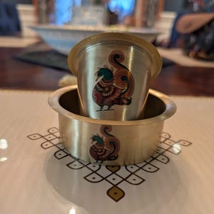 May include: A brass cup and a matching brass bowl, both featuring a colorful peacock illustration with teal, red, and orange details. The peacock design is printed on the front of each piece, showcasing a stylized bird with a curved tail.
