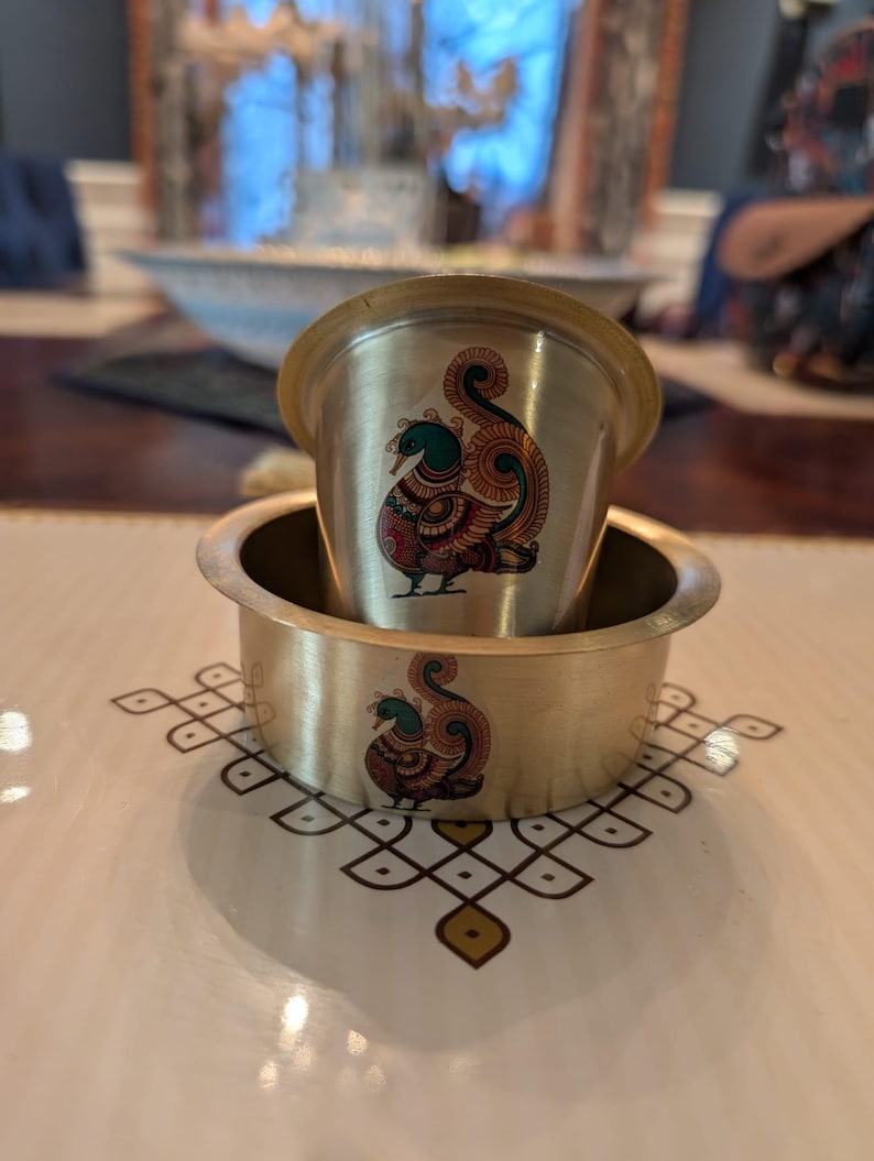May include: A brass cup and a matching brass bowl, both featuring a colorful peacock illustration with teal, red, and orange details. The peacock design is printed on the front of each piece, showcasing a stylized bird with a curved tail.