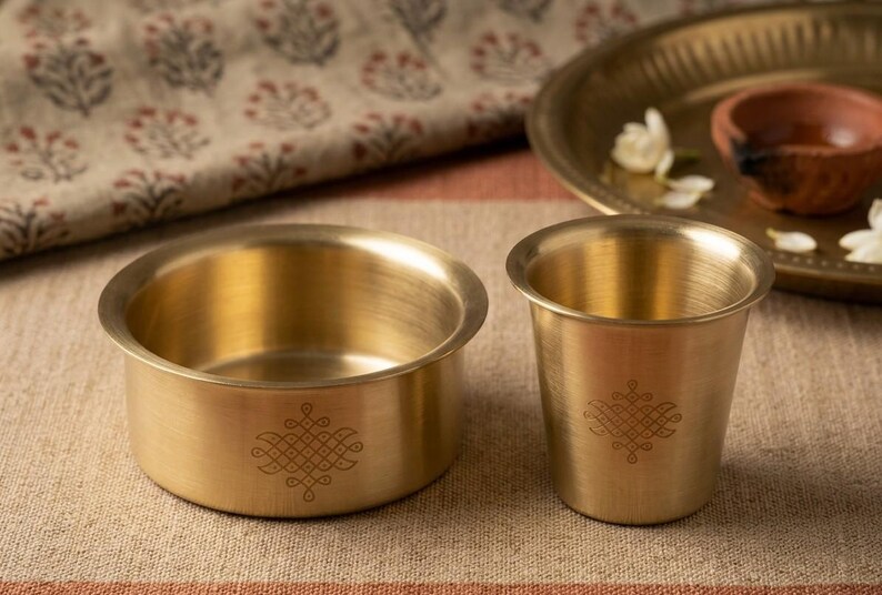 May include: A brass bowl and a matching brass tumbler, both featuring a delicate, etched geometric pattern on the exterior. The items have a smooth, metallic finish and a wide, rounded rim, showcasing a consistent design aesthetic in their construction.