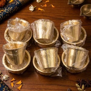 May include: A set of six brass cups and matching saucers, each individually wrapped in clear plastic. Each cup features a small, etched geometric knot design on the front. The metallic gold items are arranged in two rows of three on a dark wood surface.