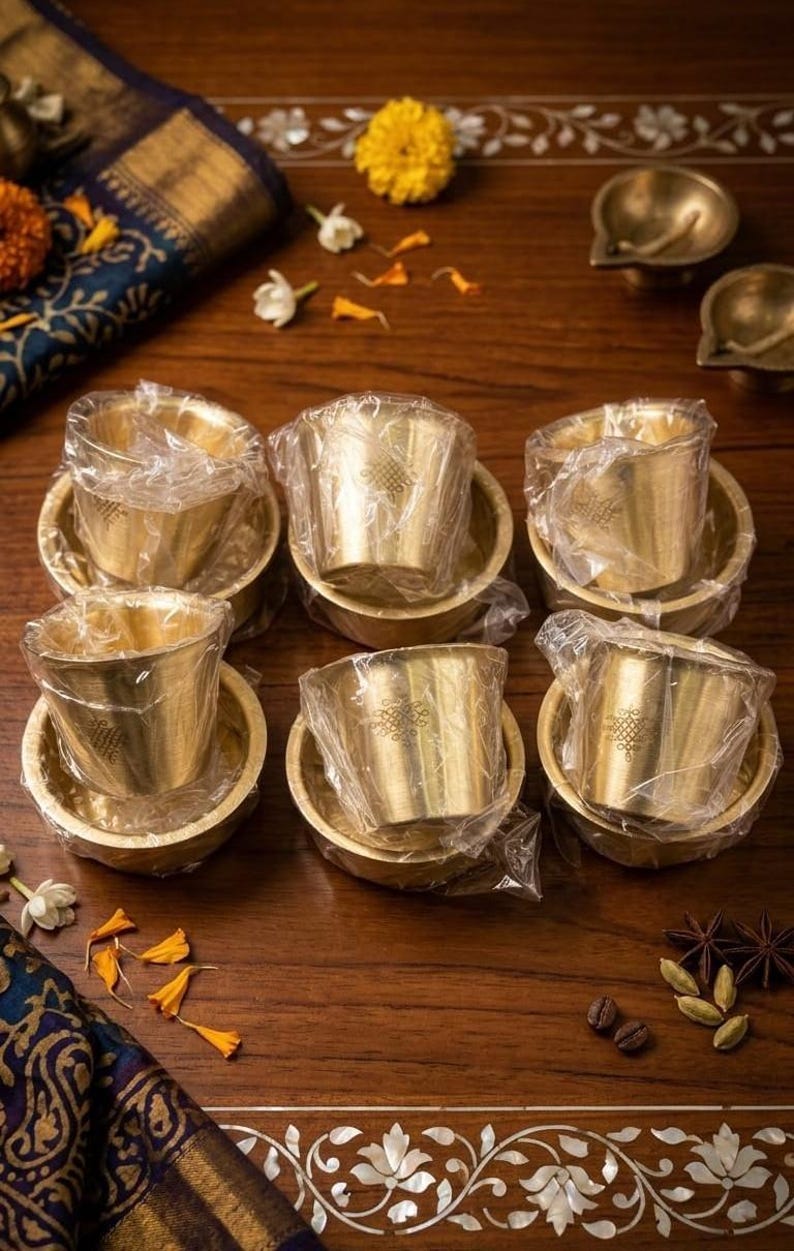 May include: A set of six brass cups and matching saucers, each individually wrapped in clear plastic. Each cup features a small, etched geometric knot design on the front. The metallic gold items are arranged in two rows of three on a dark wood surface.