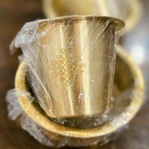 May include: A small, cylindrical brass cup with a flared rim and a decorative etched geometric pattern on the side. The cup is wrapped in clear plastic film and is positioned inside a matching brass bowl, both featuring a metallic gold finish.