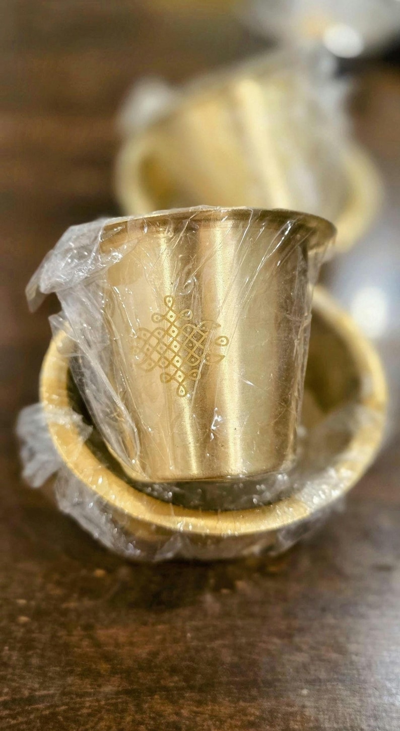 May include: A small, cylindrical brass cup with a flared rim and a decorative etched geometric pattern on the side. The cup is wrapped in clear plastic film and is positioned inside a matching brass bowl, both featuring a metallic gold finish.