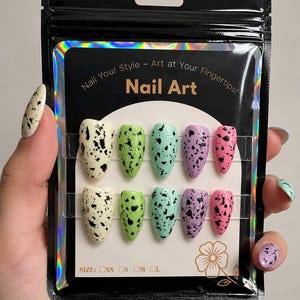 Può includere: Un set di dieci unghie finte in una confezione nera. Le unghie sono ovali e dipinte in vari colori pastello, ognuna con macchie nere. La confezione recita "Nail Your Style - Art at Your Fingertips! Nail Art."