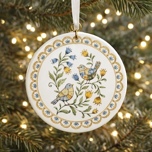 Op de afbeelding: Een rond wit keramieken ornament met twee vogels op bladerrijke takken met blauwe en gele bloemen. Het ornament heeft een decoratieve geschulpte rand in goud en blauw en hangt aan een wit lint bevestigd aan een gouden metalen ring.