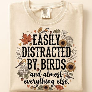 May include: A beige t-shirt featuring the black text EASILY DISTRACTED BY BIRDS and almost everything else. The design is surrounded by illustrations of colorful autumn leaves, sunflowers, white hydrangea flowers, acorns, and small birds.