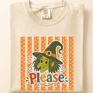 May include: A beige crew neck t-shirt featuring a graphic print of a green-skinned witch wearing a dark green hat with an orange band. Below the witch, the word Please is printed in colorful letters. The background consists of orange and white stripes.