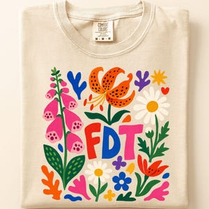 May include: A beige t-shirt featuring a colourful graphic print of various flowers, including pink foxgloves, an orange tiger lily, and white daisies, surrounding the bold, multi-coloured letters FDT in the centre of the design.