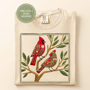 May include: Cream-colored t-shirt with an embroidered design of two cardinals perched on a branch. The birds are red and green, with floral patterns. The text "COMES WITH 4 DESIGN VARIATIONS" is in a green circle.