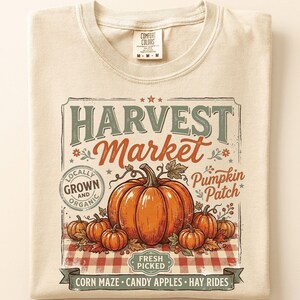 May include: A beige t-shirt featuring a graphic print with the text HARVEST MARKET, Pumpkin Patch, LOCALLY GROWN AND ORGANIC, FRESH PICKED, CORN MAZE, CANDY APPLES, and HAY RIDES. The design includes illustrations of orange pumpkins on a red checkered base.