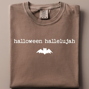 May include: A brown short-sleeved t-shirt featuring the white text "halloween hallelujah" printed in a typewriter-style font above a white graphic of a bat. The shirt has a crew neckline and a label at the inner collar reading "Comfort Colors".