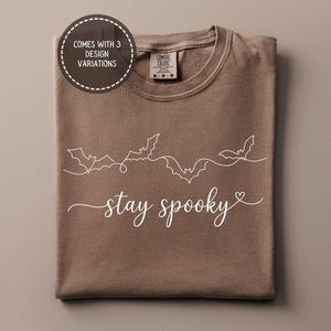 May include: A brown t-shirt featuring a white line art graphic of three flying bats connected by a continuous wavy line above the cursive text "stay spooky" with a small heart at the end. A circular badge states "COMES WITH 3 DESIGN VARIATIONS".