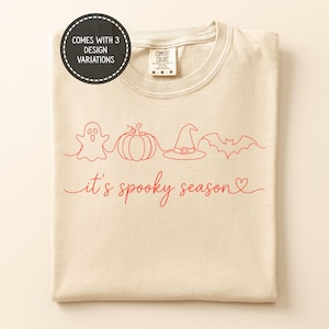 Puede incluir: Una camiseta beige con un gráfico de líneas rojas de un fantasma, una calabaza, un sombrero de bruja y un murciélago. Debajo de los iconos, el texto "it's spooky season" aparece en cursiva con un corazón. Una insignia circular indica "comes with 3 design variations".