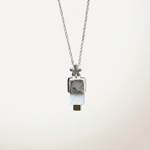 May include: A silver necklace with a pendant. The pendant has a square top, a white rectangular middle, and a small gold-coloured bottom. The chain is delicate, and the top of the pendant has a small flower design.