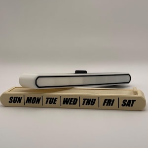 May include: Two rectangular desk organizers. The bottom one is beige with the days of the week printed in black. The top organizer is white with a black accent and a black button.