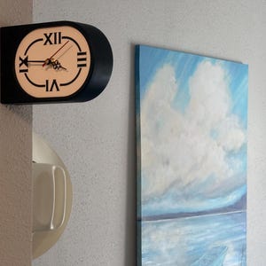 May include: A black, oval-shaped wall clock with a white face, black Roman numerals, and thin black hands. The clock is mounted on a corner. Beside it is a vertical rectangular painting of a blue sky with white clouds over a body of water.