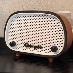 May include: A retro-style speaker with a white perforated front, brown sides, and black accents. The speaker has two black knobs and the Google logo. It sits on a dark surface.