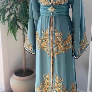 May include: A floor-length teal kaftan dress featuring intricate gold embroidery with sequins and beads. The garment has long, wide sleeves, a V-neckline, and a wide matching waistband secured with a decorative gold-toned brooch at the center.