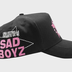 Op de afbeelding: Zwarte baseballcap met een gebogen klep. De cap heeft de tekst "SAD BOYZ" in roze, omlijnd door een witte ster. Boven de ster staat de tekst "UNA ESTRELLA GIGANTE SE VE" in het wit.
