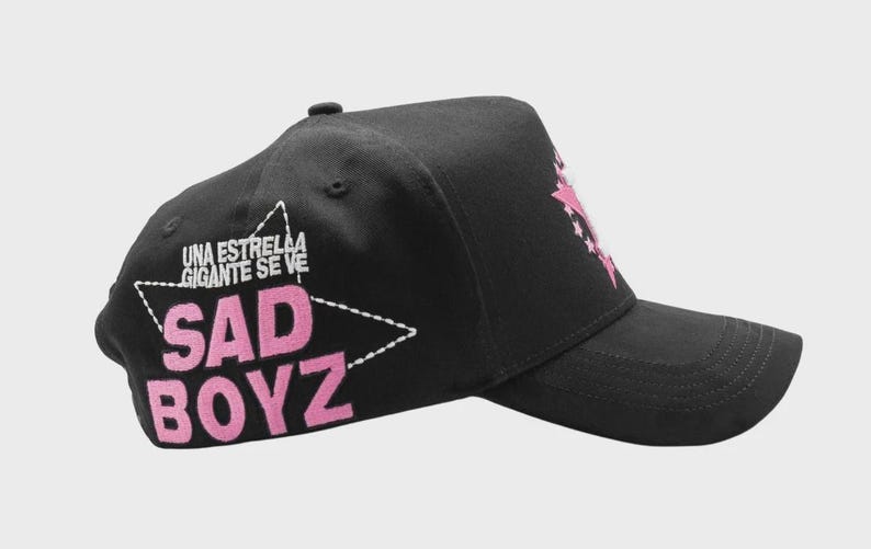 Op de afbeelding: Zwarte baseballcap met een gebogen klep. De cap heeft de tekst "SAD BOYZ" in roze, omlijnd door een witte ster. Boven de ster staat de tekst "UNA ESTRELLA GIGANTE SE VE" in het wit.