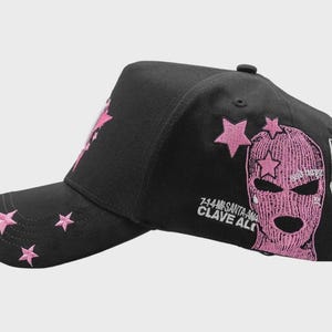 Op de afbeelding: Zwarte baseballcap met roze sterrenborduurwerk. De cap heeft een roze bivakmuts en de tekst "7-14-MS SANTA-ANA CLAVE ALTO" en een afbeelding van een klaptelefoon.