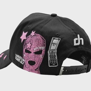 Op de afbeelding: Zwarte baseballcap met roze borduursels. Het heeft een roze bivakmuts-afbeelding met sterren, een klaptelefoon en de tekst "CLAVE ALTO". De achterkant heeft een wit "dh"-logo en een verstelbare band.